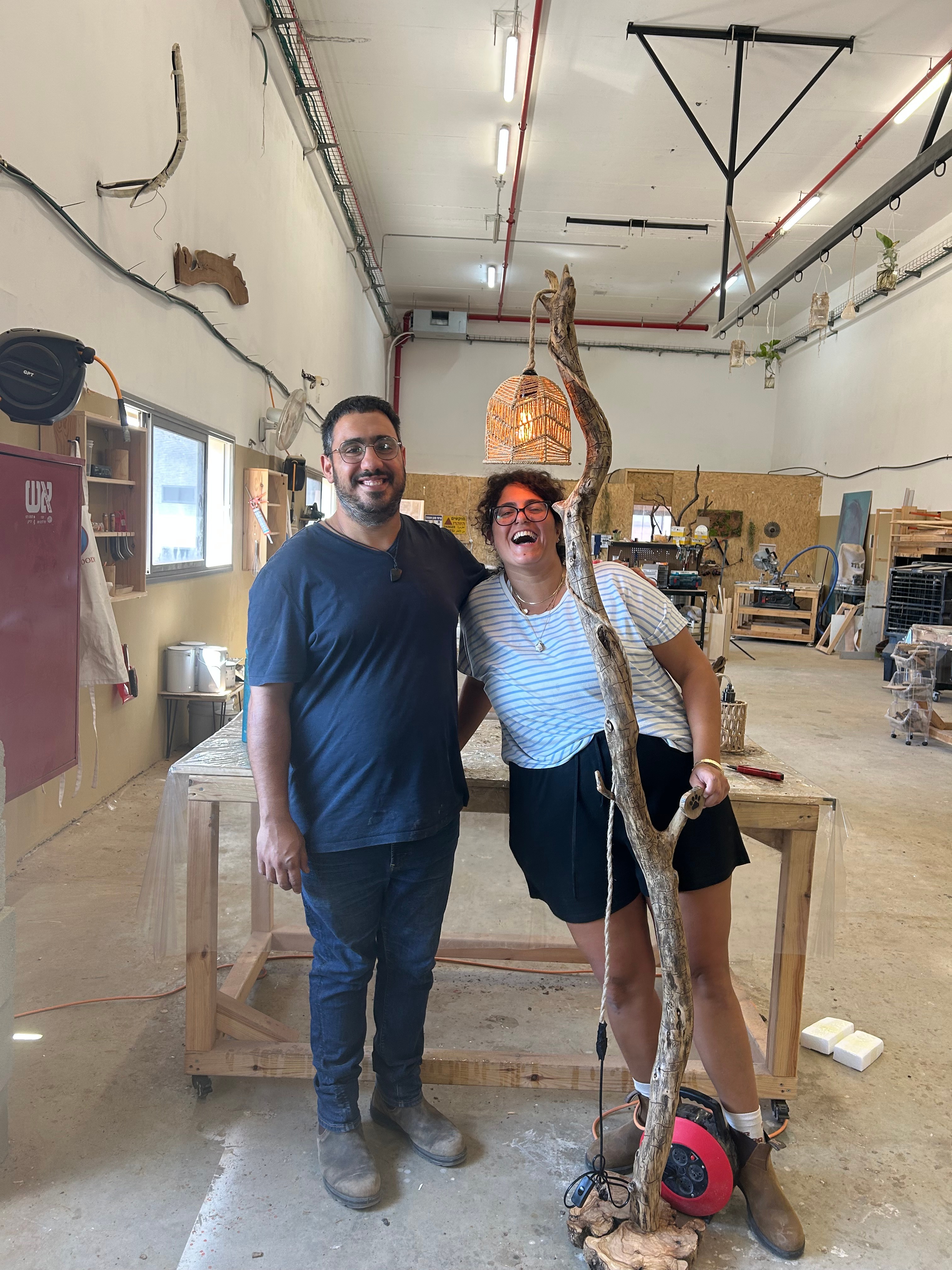 זוג יוצרים מנורת רצפה מענף עץ טבעי עם אהיל קלוע בסדנת נגרות